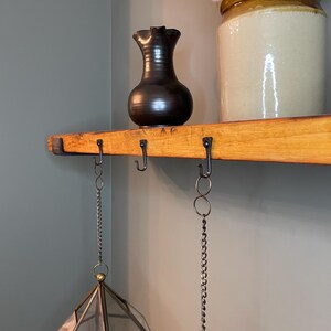 May include: A wooden shelf with two hanging lanterns. The lanterns are made of glass and have a geometric shape. The shelf has black metal hooks and chains. A brown ceramic pitcher and a white ceramic jar with a brown lid are on the shelf.