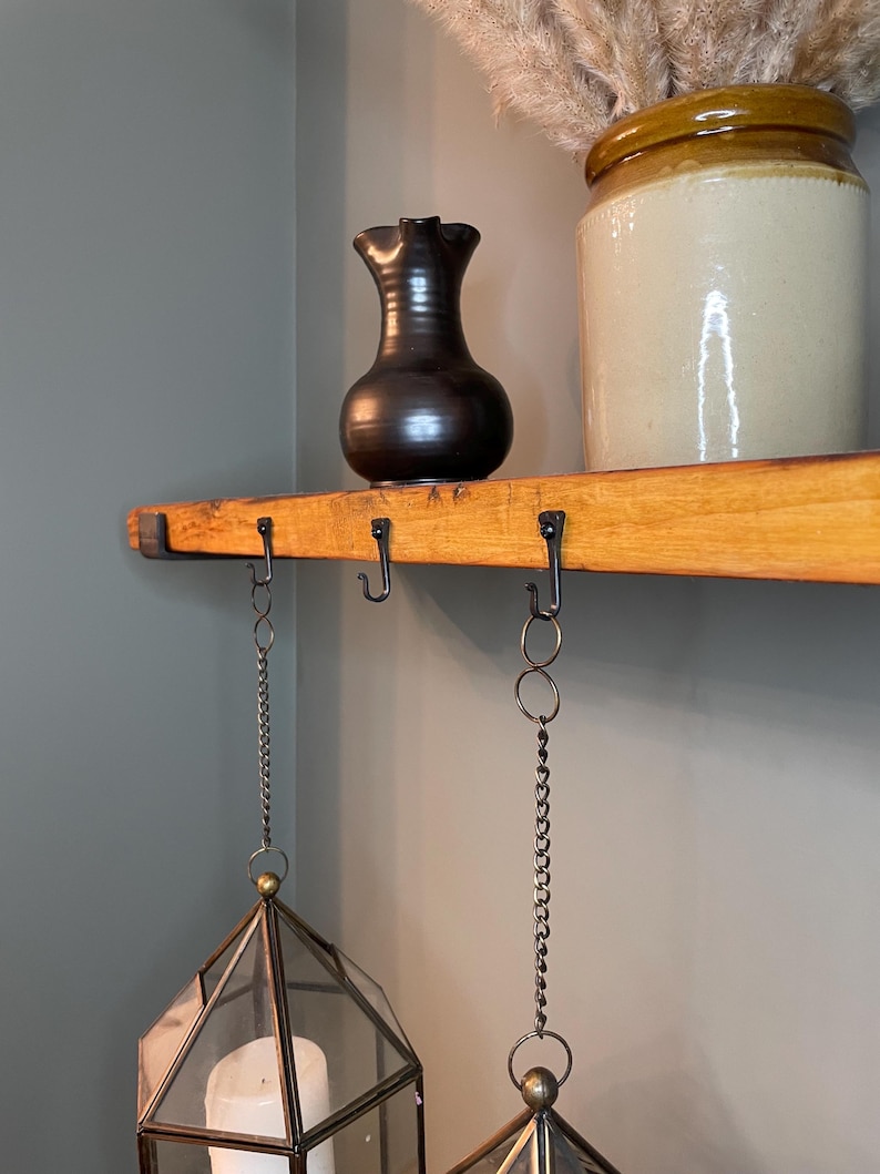 May include: A wooden shelf with two hanging lanterns. The lanterns are made of glass and have a geometric shape. The shelf has black metal hooks and chains. A brown ceramic pitcher and a white ceramic jar with a brown lid are on the shelf.