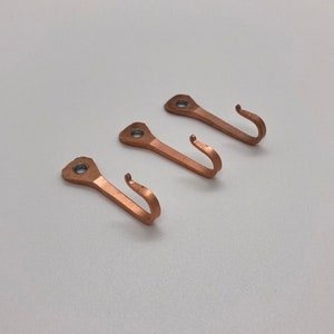 May include: Three copper hooks with a simple design. The hooks are all the same size and shape, and they have a slightly rounded top. They are perfect for hanging keys, coats, or other small items.