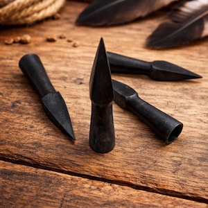Puede incluir: Primer plano de cuatro puntas de flecha negras con puntas cónicas y afiladas, dispuestas sobre una superficie de madera rústica. Las puntas de flecha son de formas y tamaños variados. Plumas y cuerda en el fondo.