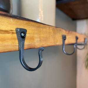 Könnte beinhalten: Eine Wandgarderobe bestehend aus einem hellbraunen Naturholzbrett mit Baumkante. Drei schwarze Metallhaken sind mit schwarzen Schrauben an der Vorderseite des Holzes befestigt. Die Haken haben ein geschwungenes Design mit einer kleinen Zierschnörkel.