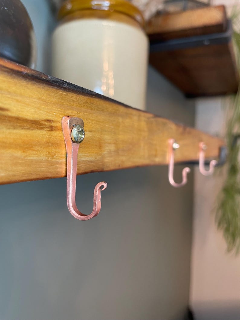 May include: Three copper hooks are screwed into a wooden shelf. The hooks are curved and have a shiny finish.