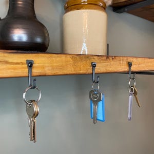 May include: A wooden shelf with three hooks holding keys. The shelf is made of natural wood and has a rustic look. The hooks are black metal and are attached to the underside of the shelf. The keys are silver and have different shapes and sizes.