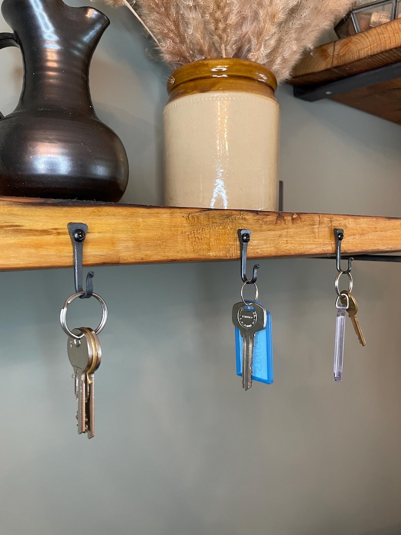 May include: A wooden shelf with three hooks holding keys. The shelf is made of natural wood and has a rustic look. The hooks are black metal and are attached to the underside of the shelf. The keys are silver and have different shapes and sizes.