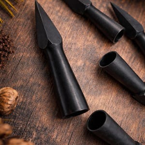 May include: Close-up of several black metal spearheads with a conical tip and cylindrical base. The spearheads are arranged on a wooden surface, with a few pine needles and a pine cone in the background. The spearheads are likely for hunting or display.