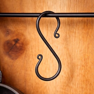 May include: A matte black metal S-hook hanging from a thin horizontal rod. The hook features a smooth, curved design with rounded, curled ends at both the top and bottom, creating a distinct S-shape profile for hanging various household items.