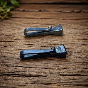 May include: Two dark grey, hand-forged metal key hooks with a folded, J-shaped design. Each hook features a flat, rectangular head stamped with a small, distinct mark. The metal has a textured, matte finish, showcasing the craftsmanship of the iron.