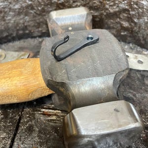 May include: A small, black, hand-forged metal hook with a curled end and a single circular mounting hole, resting on the flat, textured steel head of a heavy-duty hammer with a wooden handle.