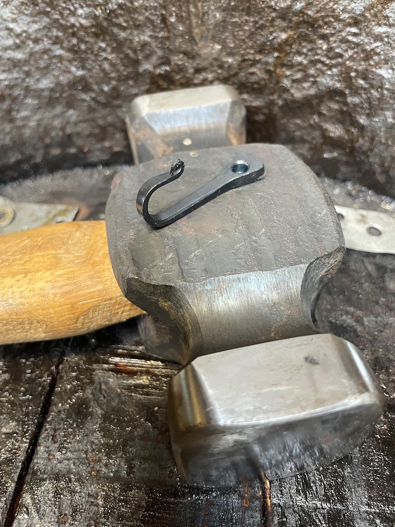May include: A small, black, hand-forged metal hook with a curled end and a single circular mounting hole, resting on the flat, textured steel head of a heavy-duty hammer with a wooden handle.