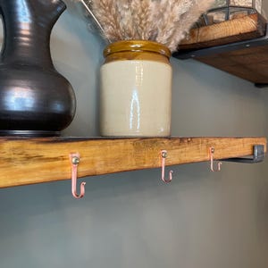 May include: A wooden shelf with three copper hooks attached to the underside. The shelf is stained a light brown color and has a rustic appearance. A ceramic vase with dried pampas grass sits on the shelf.