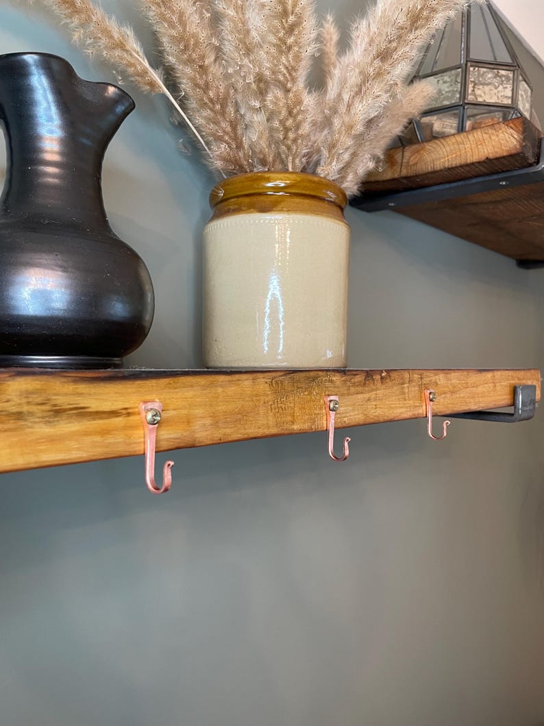 May include: A wooden shelf with three copper hooks attached to the underside. The shelf is stained a light brown color and has a rustic appearance. A ceramic vase with dried pampas grass sits on the shelf.