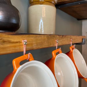 May include: Three orange ceramic pots with white interiors hang from a wooden shelf with copper hooks. The pots have handles and are ready to be used for cooking.