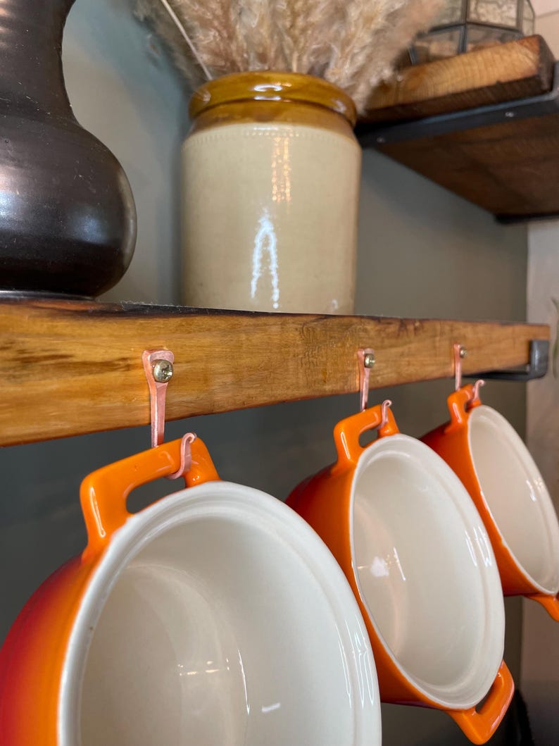 May include: Three orange ceramic pots with white interiors hang from a wooden shelf with copper hooks. The pots have handles and are ready to be used for cooking.