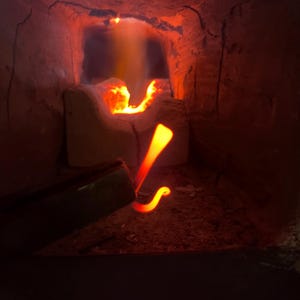 May include: A close-up of a metal rod being heated in a forge. The rod is glowing orange-red and is bent into a hook shape. The forge is made of brick and has a small opening where the flames are visible.