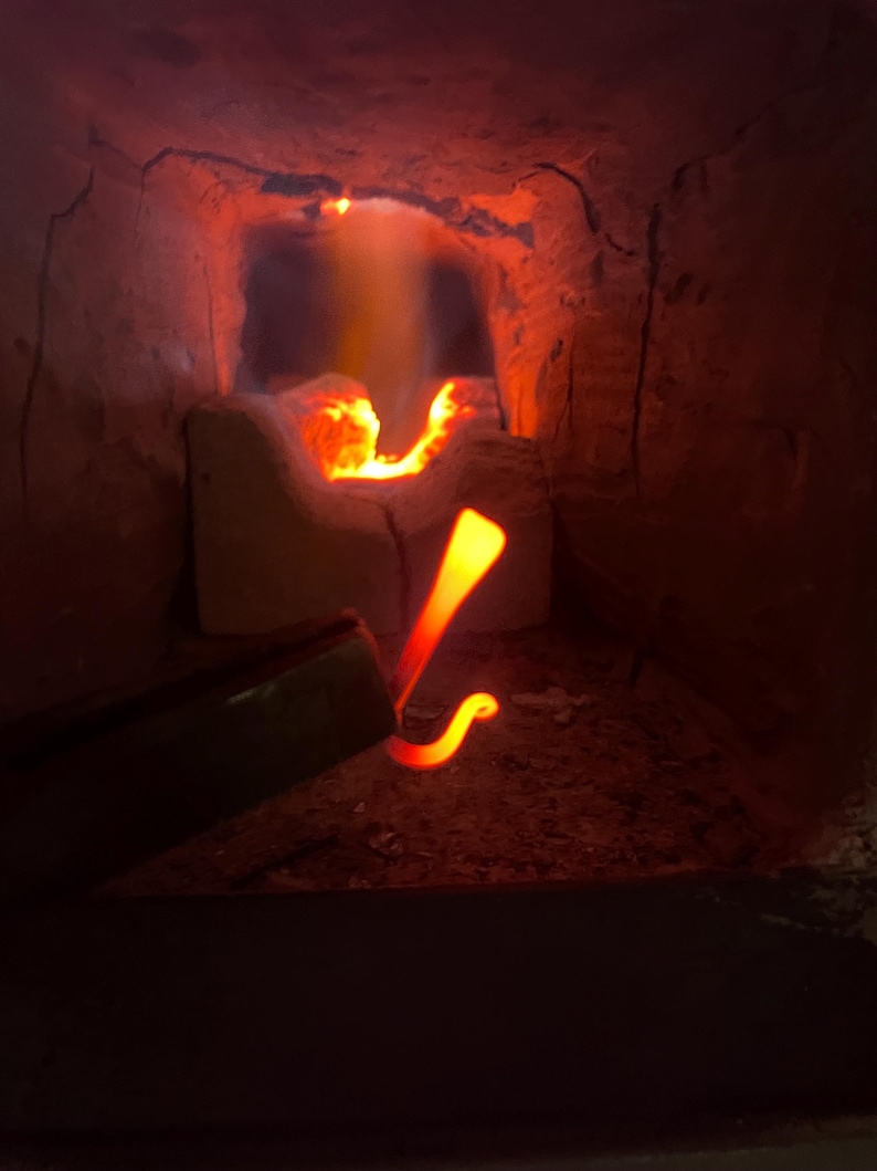 May include: A close-up of a metal rod being heated in a forge. The rod is glowing orange-red and is bent into a hook shape. The forge is made of brick and has a small opening where the flames are visible.