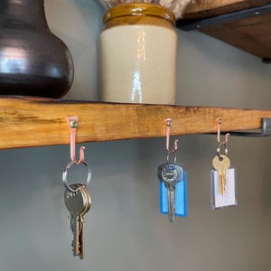 May include: Three sets of keys hanging on copper hooks mounted on a wooden shelf. The keys are attached to key rings and have different colored key tags. The shelf is made of wood and has a rustic finish.