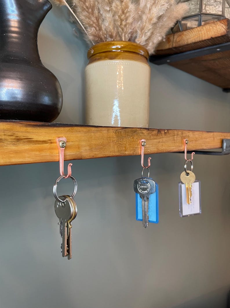 May include: Three sets of keys hanging on copper hooks mounted on a wooden shelf. The keys are attached to key rings and have different colored key tags. The shelf is made of wood and has a rustic finish.