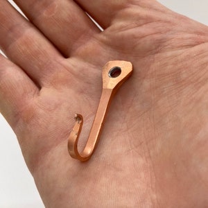 May include: A small, copper-colored hook with a rounded top and a hole in the center. The hook is bent into a J-shape.