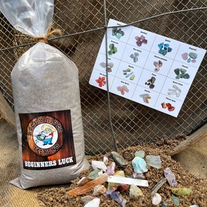 May include: A bag of dirt labelled "Beginners Luck" from Rocky River Mining. The bag is sitting on a hessian surface next to a pile of colourful gemstones and a chart with names of different types of gemstones.