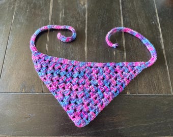 May include: A triangular crochet bandana featuring a variegated yarn pattern in shades of bright pink, purple, and blue. The accessory is constructed with an open-weave stitch and includes two thin crocheted ties extending from the top corners.