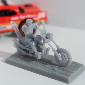 May include: A gray 3D printed model of a motorcycle with a rider. The motorcycle has a long front fork and a single seat. The rider is wearing a helmet and is holding the handlebars.