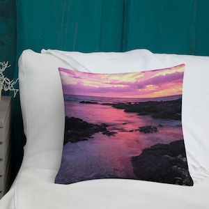 Può includere: Cuscino quadrato decorativo con una scena di tramonto su una costa rocciosa. L'immagine mostra un gradiente di tonalità rosa, viola e arancioni nel cielo e nell'acqua, creando un'estetica serena e colorata.