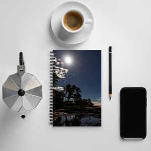Può includere: Un quaderno a spirale con una copertina nera che presenta una fotografia di un cielo notturno con luna piena e palme riflesse nell'acqua.