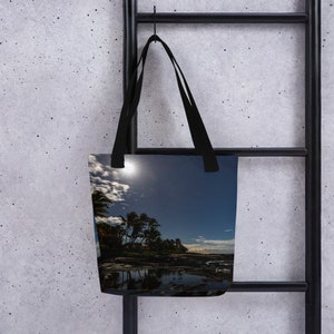 Può includere: Una borsa tote nera con una luna piena e palme riflesse nell'acqua di notte. L'immagine è una fotografia di una scena di spiaggia con un cielo stellato.