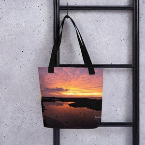 Può includere: Una borsa tote con un tramonto riflesso nell'acqua. Il cielo è di un arancione e rosa vibrante con le nuvole. L'acqua è di un blu scuro con un riflesso del tramonto.