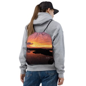 Può includere: Una borsa a sacco con un tramonto riflesso nell'acqua. La borsa è realizzata in un tessuto morbido e resistente e presenta un tramonto vibrante arancione, rosa e viola con una sagoma scura di una costa rocciosa. La borsa ha un cordoncino nero e una firma bianca "Sun More" nell'angolo in basso a destra.