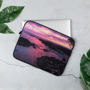 Può includere: Una custodia per laptop con un tramonto colorato su una spiaggia rocciosa. Il cielo è un mix di rosa, viola e arancione. L'acqua è di un blu profondo e le rocce sono di colore grigio scuro. La custodia è realizzata in un materiale morbido e resistente e ha una chiusura a zip.