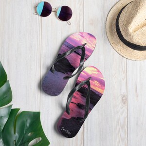 Può includere: Un paio di infradito con un design tramonto, con sfumature di rosa, viola e arancione. Le infradito hanno cinturini neri e le parole "Sara More" stampate sulla suola. Sono visibili anche occhiali da sole e un cappello di paglia.