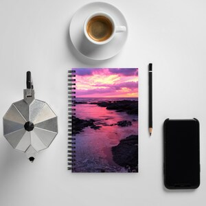 Può includere: Un quaderno a spirale con un tramonto colorato sull'oceano sulla copertina. Il quaderno è su una superficie bianca con una tazza di caffè, una matita e uno smartphone.