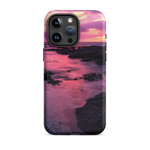 Può includere: Un tramonto rosa e viola su una costa rocciosa con onde che si infrangono sulla riva. L'immagine è stampata su una custodia per telefono nera con un ritaglio per l'obiettivo della fotocamera.