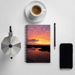 Può includere: Un taccuino a spirale con un'immagine di tramonto sulla copertina. Il cielo infuocato si riflette nell'acqua calma. Una tazza di caffè, una matita, un telefono e una caffettiera sono visibili.