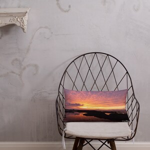 Può includere: Un cuscino decorativo con una fotografia di un tramonto su uno specchio d'acqua. Il cielo è un mix vibrante di rosa, arancione e viola. L'acqua riflette i colori del cielo. Il cuscino è seduto su una sedia con un cuscino bianco.