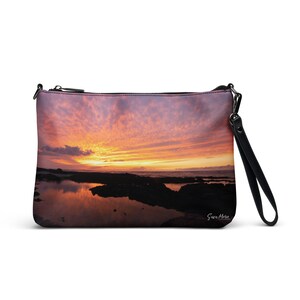 Può includere: Una pochette nera con cerniera con un tramonto sull'oceano riflesso in una pozza di marea stampato sul davanti. La borsa ha una tracolla nera.