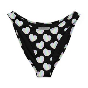 May include: Black bikini bottoms with a repeating pattern of iridescent heart shapes. The hearts have a subtle rainbow sheen. The brand name "THE MULLEIN" is printed on the back of the garment.