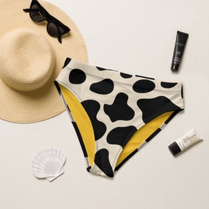 May include: Black and white cow print bikini bottom with a yellow lining. The bikini bottom is laying on a white surface with a straw hat, seashell, and two tubes of sunscreen.