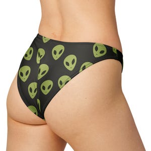 May include: Black bikini bottoms with a repeating pattern of green alien faces. The swimwear has a classic cut and is made of a smooth, stretchy fabric. The alien design adds a fun, extraterrestrial touch to the beachwear.