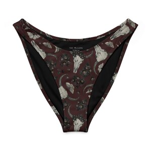 May include: High-waisted bikini bottoms with a dark burgundy background and a repeating pattern of white skulls, moths, crescent moons, and floral arrangements. The inner lining is black, and the text "THE MULLEIN TORCH" is visible.