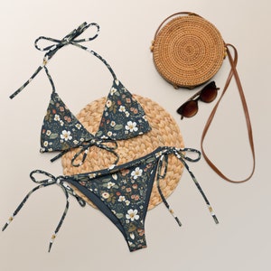 May include: A dark blue bikini with a floral print. The bikini top is a triangle style with adjustable straps. The bikini bottom is a high-cut style with ties on the sides. The bikini is laying on a woven straw mat.