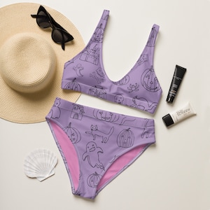 May include: Purple bikini with a black and white Halloween print featuring ghosts, pumpkins, cats, and skulls. The bikini top has a scoop neck and adjustable straps. The bikini bottom is a high-waisted style.
