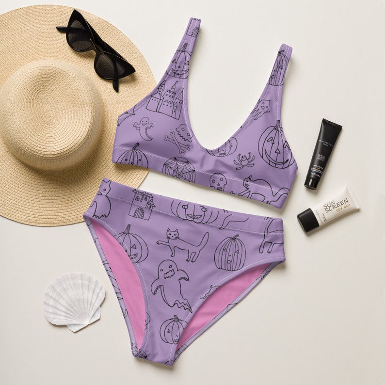 May include: Purple bikini with a black and white Halloween print featuring ghosts, pumpkins, cats, and skulls. The bikini top has a scoop neck and adjustable straps. The bikini bottom is a high-waisted style.