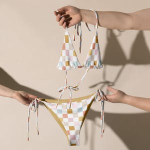 May include: A checkered bikini top and bottom with pink, yellow, and blue squares. The bikini top has adjustable straps and ties at the neck. The bikini bottom has ties on the sides.