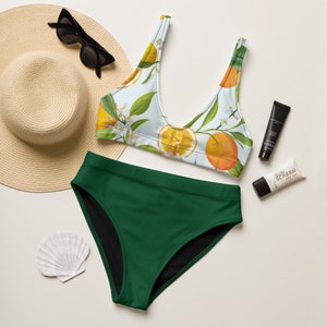 May include: A green bikini bottom with a white and orange floral print bikini top. The bikini top has a scoop neck and adjustable straps. The bikini is laid out on a white surface with a straw hat, sunglasses, and sunscreen.