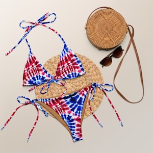 May include: A red, white, and blue tie-dye bikini with a matching top and bottom. The bikini is laid out on a woven straw mat with a round straw bag and sunglasses.