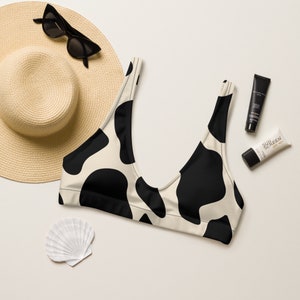 May include: A beige and black cow print bikini top. The top is paired with a straw hat, black sunglasses, sunscreen, and a seashell. The bikini top has a scoop neck and is made of a soft, stretchy material.