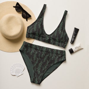 May include: A black and green swimsuit with a leafy print. The swimsuit has a halter top and high-waisted bottoms. A straw hat, sunglasses, and sunscreen are also pictured.