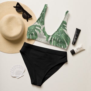 May include: A black bikini with a tropical leaf print top and solid black bottoms. A straw hat, sunglasses, and sunscreen are also pictured.